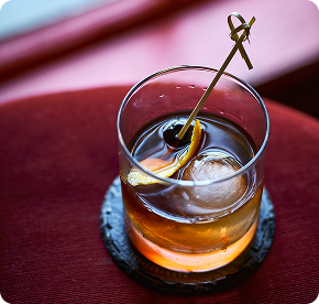 Maple bourbon cocktail in a rocks glass with a large ice sphere, garnished with an orange twist and skewered cherries, served on a black coaster against a deep red background.