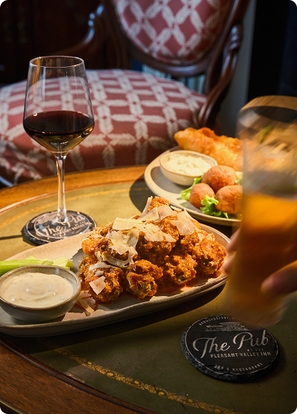 Plate of crispy wings topped with shaved parmesan, served with dipping sauce, alongside a glass of red wine on a table at The Pub at Pleasant Valley Inn.